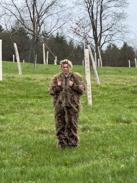 Person in a ghillie suit standing in a grassy field with young
trees protected by tree tubes.